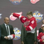 Dennis Cholowski, center, puts on his sweater with members of the Detroit Red Wings management team and others at the NHL draft in Buffalo, N.Y., Friday June 24, 2016. (Nathan Denette/The Canadian Press via AP) MANDATORY CREDIT