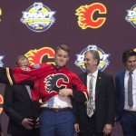 Matthew Tkachuk, center, sixth overall pick, pulls on his sweater as he stands on stage with members of the Calgary Flames management team and others at the NHL draft in Buffalo, N.Y., Friday June 24, 2016. (Nathan Denette/The Canadian Press via AP) MANDATORY CREDIT