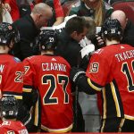 Calgary Flames center Sean Monahan (23), center Austin Czarnik (27) and left wing Matthew Tkachuk (19) look on as training staff treat head coach Bill Peters who was struck in the face with a puck during the second period of an NHL hockey game against the Arizona Coyotes, Sunday, Nov. 25, 2018, in Glendale, Ariz. (AP Photo/Ross D. Franklin)