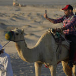 A man takes a selfie while riding camels in Mesaieed, Qatar, Nov. 26, 2022. Throngs of World Cup fans in Qatar looking for something to do between games are leaving Doha for a classic Gulf tourist experience: riding a camel in the desert. But the sudden rise in tourists is putting pressure on the animals, who have almost no time to rest between each ride. (AP Photo/Ashley Landis)