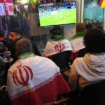 
              Lebanese soccer fans of Iran's team cover their backs with Iranian flags, as they sit at a coffee shop smoking water pipes and watch the World Cup group B soccer match between Iran and the United States, in the Hezbollah stronghold in the southern suburbs of Beirut, Lebanon, Tuesday, Nov. 29, 2022. (AP Photo/Hussein Malla)
            