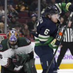 Arizona Coyotes goalie Karel Vejmelka, left, of the Czech Republic, is struck in the mask by the puck as Vancouver Canucks' Brock Boeser stands in front of him during the first period of an NHL hockey game in Vancouver, British Columbia on Saturday, Dec. 3, 2022. (Darryl Dyck/The Canadian Press via AP)