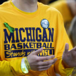 A student wears a green ribbon and a Michigan State button on his Michigan basketball T-Shirt before an NCAA college basketball game between Michigan and Michigan State, Saturday, Feb. 18, 2023, in Ann Arbor, Mich. The university is honoring the victims of the Michigan State University shooting that killed three and injured five Monday. (AP Photo/Carlos Osorio)