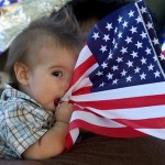 Eli Baldonado, 7-months-old, grabs hold of a nearby flag on Thursday, July 3, 2008, at Apodaca Park in Las Cruces, N.M., as his grandfather, Isidro Salcedo holds him. Salcedo was helping build the Kids Kindness Day float for the city's annual Electric Light Parade held to celebrate Independence Day. (AP Photo/Las Cruces Sun-News, Norm Dettlaff)