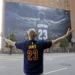 
              FILE - In this June 9, 2015, file photo, a fan poses for a photo in front of a billboard featuring Cleveland Cavaliers forward LeBron James before Game 3 of basketball's NBA Finals in Cleveland. Two people familiar with the negotiations say LeBron James has agreed to a one-year, $23 million contract with the Cavaliers for next season. The deal includes a player option for 2016-17. The people spoke to The Associated Press on condition of anonymity Thursday because the contract has not been signed. James has informed the team he will return. (AP Photo/Tony Dejak)
            